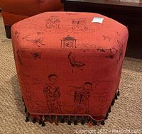 Full view of hexagonal reddish-orange foot rest with black detailed chinoiserie print and tassel bead trim on bottom.