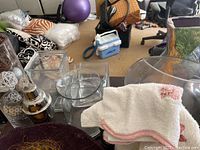 Glass vases, serving bowl, decorative filler balls, and white cloth on table with background of room and various items.