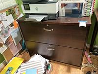 Front and side view of the large wooden filing cabinet with two drawers, showing metal handles and key locks, top surface holding printer and file folders.