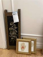 Three piece lot showing two small gold framed prints leaning on the wall in front of a wall corner and next to a medium vertical rectangular vintage metal daisy art piece with embossed floral frame.