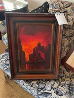 Front view of framed painting showing cityscape silhouette against fiery sky in wooden frame with decorative inner border.