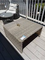 Large square outdoor wicker table with glass top on a deck, showing wear and placed next to two wicker chairs with cushions.