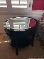 Full front view of round dark wooden side table with glass inset top, placed next to a window and a sofa.