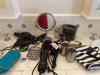 Overview of hair dryers, hot air brush, makeup mirror, metal soap dish, tissue box holder, stainless cups, toiletry container, cosmetics bag on bathroom counter