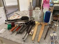 Photo showing a collection of kitchen utensils including wooden spatulas, metal scoops, collectible spoons, olive oil and vinegar containers, tongs, and other utensils arranged on a marble countertop.
