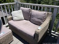 Outdoor couch on wooden deck with beige woven synthetic frame and light purple seat cushions, visible wear and stains.