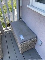 Rectangular wicker outdoor ottoman with cushion, placed on a balcony deck beside exterior wall and railing.