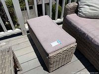 Top view of rectangular outdoor ottoman with purple cushion on beige woven wicker base, showing wear and dirt on cushion.
