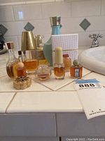 Wide shot showing entire assortment of nine vintage perfume bottles on white tiled surface by bathroom sink with varying shapes, sizes and colors.