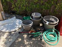 Overview of round foldable white table top, gray gardening pots in multiple sizes, green garden hose, and assorted gardening tools on driveway surface.