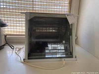 Front view of the Ronco Showtime Rotisserie BBQ showing the entire unit with power cord on a white surface near window with blinds.