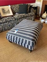 Front side view of the striped upholstered ottoman showing its rectangular shape and caster wheels.