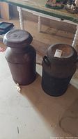 Two metal antique milk jugs shown side by side on concrete floor with table in background.