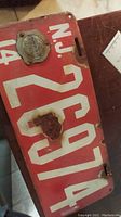 Photo showing full view of 1914 NJ license plate with attached round certified maker's plaque and visible rust spots and wear.