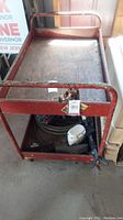 Photo showing red metal rolling utility cart with two shelves, paint chipping and weathered condition.