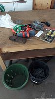 Photo showing green plastic basket, black bucket with chain, multiple tie-down straps, safety goggles, and tie down kit on wood table.