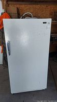 Front view of white Estate stand up freezer with a handle on the left side in a garage setting.