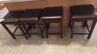 Four brown kitchen stools lined up against a wooden panel wall on a tiled floor.