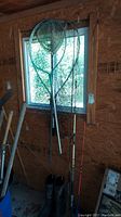 Two fishing nets, two fishing rods, and a pair of men's black rubber boots arranged leaning against a windowed wall in a garage.