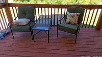 Wide shot showing two cushioned chairs and glass top table on a wooden deck with pillows.