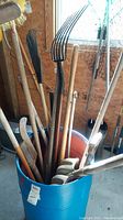 Blue barrel filled with assorted garden tools including shovels, pitchforks, rakes with wooden handles, showing metal working heads and handles varying in length.