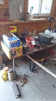 Photo showing a table with multiple items including boxes, sprayers, a red spreader funnel, various hand tools, and part of a wheelbarrow.