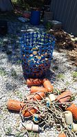 Orange basket with circular holes standing upright on gravel in outdoor setting, several orange and white cylindrical water markers attached to ropes lying in front of basket.