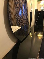 Close side view of large attached oval mirror mounted on black lacquer dresser surface reflecting patterned curtains and room environment.
