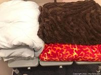 Three blankets folded and displayed: white quilted, brown faux fur, and red with orange pattern.