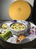 View of the open round yellow box containing six decorative coffee cups arranged on a white satin-like liner. Cups display various colorful butterfly designs.