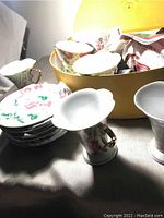 Twelve decorated cups and saucers stacked with a yellow box in background