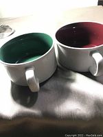 Two white ceramic soup mugs with green and dark red interiors side by side on fabric surface.
