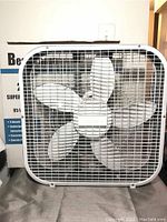 Front view of white 20 inch box fan, showing metal grille and 5 white blades. Original product box is visible behind the fan.