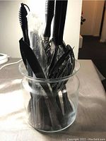 Photo of multiple black handled kitchen tools stored in a glass jar. Items include knives, forks, and peelers.