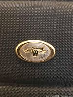 Close-up photo of metallic oval brand emblem with 'W' and wings logo on luggage surface