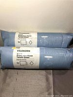 Two blue IKEA VILDKORN Queen size pillows in unopened packaging laid horizontally on gray surface against a light gray wall.
