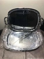 Photo showing large silver-plated serving tray with ornate handles and decorative edges, standing upright behind two trays laying flat wrapped in plastic.