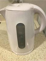White Mainstay electric water kettle with handle, spout, and water level window on kitchen counter.