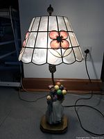 The lamp is shown illuminated on a floor with a white shell panel lampshade decorated with pink flower petals and a scalloped lower edge.