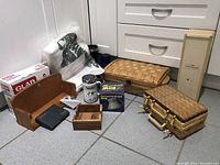 Wide shot showing all items included in the lot arranged on floor against cabinetry. Items include two woven baskets, wooden holders, mugs, blue glasses, plastic wrap and boxed wine opener.