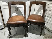 Pair of two 1940's office chairs with brown leather upholstery and carved wooden frames, showing front and side views in a kitchen corner.
