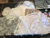 Photo showing several large and medium white crochet doilies and one square embroidered needlework piece with floral design and red border on a table.