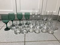 Front row includes green tall goblets, clear short tumblers, and assorted clear glasses behind them, all arranged on a tiled floor.