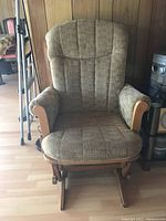 Front view of brown fabric glider rocking chair with wooden armrests and base on light wood floor.