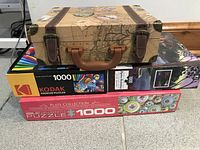 Stack of three 1000-piece jigsaw puzzles including one in a decorative travel case box with map and leather strap design, a Kodak hot air balloons puzzle, and a plate collection graphic puzzle.