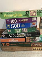 Stack of seven puzzle boxes showing titles: Disney Snow White, Jumanji, Guild 500-piece, Windsor 500-piece, Pusheen cat tea party, Brewed Awakenings, and Anne Geddes