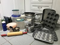 Photo of assorted kitchenware items on a tiled floor including bakeware, pots, plastic containers, and utensils.