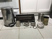 View of all stainless steel kitchen accessories including a pedal bin, small cups, sugar bowls, lidded container, cylindrical holders, and a bread box.