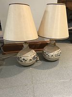 Pair of ceramic table lamps with beige shades and blue decorative patterns on bases, placed on tiled floor beneath a mirror, showing crack detail and overall condition.