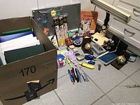 Wide view of lot showing green and white binders in cardboard box, microscope, small toys, pens, books, and a bulletin board on floor.